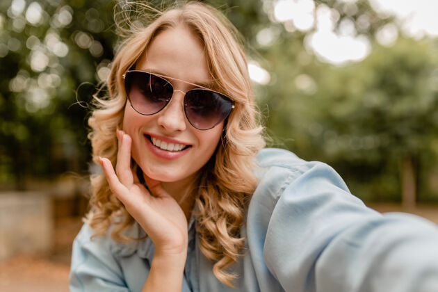 微笑迷人的金发微笑的白牙女人穿着夏装在公园里散步在电话里自拍时尚模特明亮