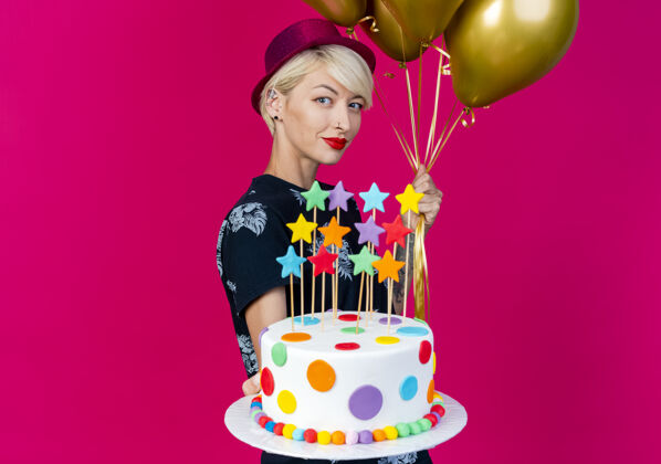 复制高兴的年轻金发派对女孩戴着派对帽站在侧视图拿着气球和伸展生日蛋糕与星星对着摄像机在深红色背景与复制空间隔离蛋糕走向气球