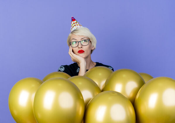无聊无聊的金发派对女郎戴着眼镜和生日帽站在气球后面把手放在下巴下看着紫色背景上孤立的一面帽子女孩下巴