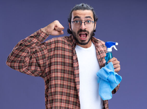 小伙子兴奋的年轻帅气的清洁工穿着t恤 拿着抹布喷雾瓶 在蓝色的墙上显示“是”的手势瓶子喷雾抹布