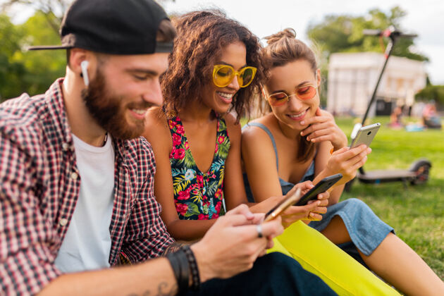 兴奋快乐的年轻朋友们坐在公园里用智能手机微笑 男人和女人一起玩友谊无线朋友