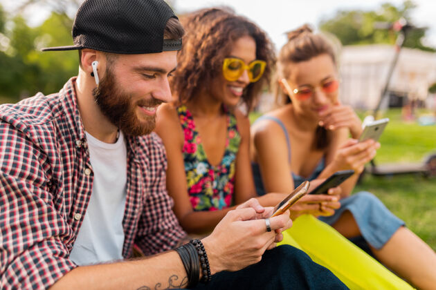 女人快乐的年轻朋友们坐在公园里用智能手机微笑 男人和女人一起玩耳机电话时尚
