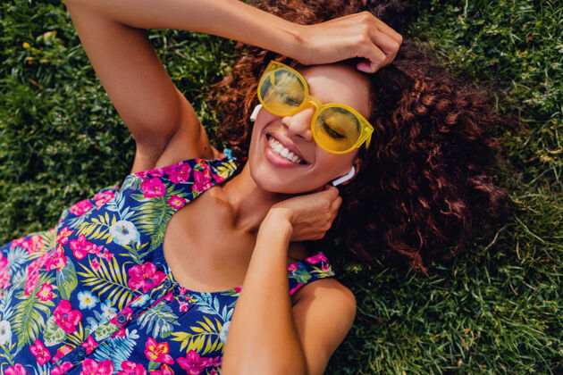 女人年轻时尚的黑人女子 戴着无线耳机听音乐 躺在公园的草地上玩得很开心 夏日的时尚风格 五颜六色的时髦装扮 俯瞰风景女性耳机耳机