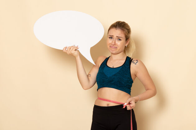 量正面图身穿运动服的年轻女性手持白色标志 在白色墙上测量身体健康运动美容运动适合健身身体腹
