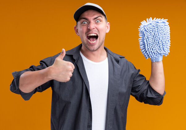 破布兴奋的年轻帅气的清洁工穿着t恤 戴着帽子 拿着抹布 在橙色的墙上孤立地竖起大拇指家伙抱着清洁