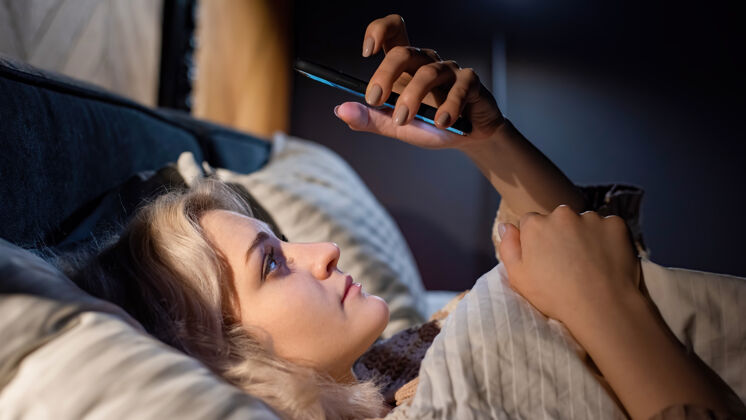 卧室年轻的金发女孩在床上用她的智能手机试图入睡社交媒体成瘾年轻女孩女人