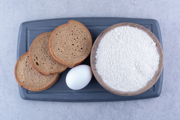 面粉一小碗面粉 一个鸡蛋和三片面包放在大理石表面的海军蓝木板上饮食碗鸡蛋
