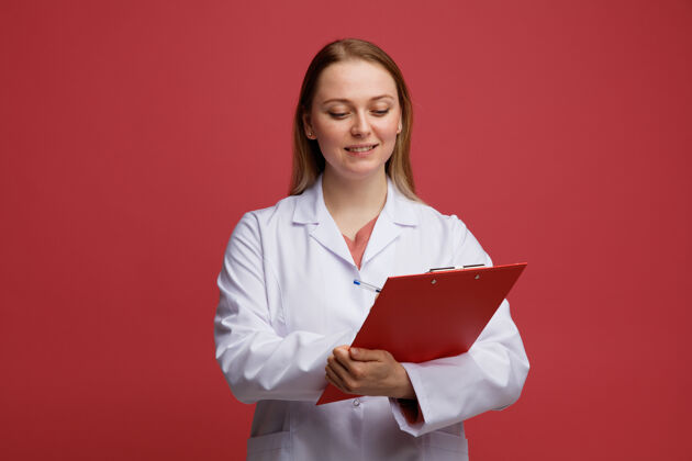 女性微笑着的年轻金发女医生 穿着医用长袍 脖子上戴着听诊器 在写字板上用笔写字粉红色周围钢笔