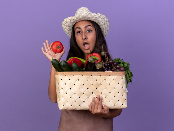 惊喜年轻的园丁女孩围着围裙 戴着夏帽 手里拿着装满蔬菜和新鲜番茄的板条箱 站在紫色的墙上 既高兴又惊讶围裙蔬菜西红柿