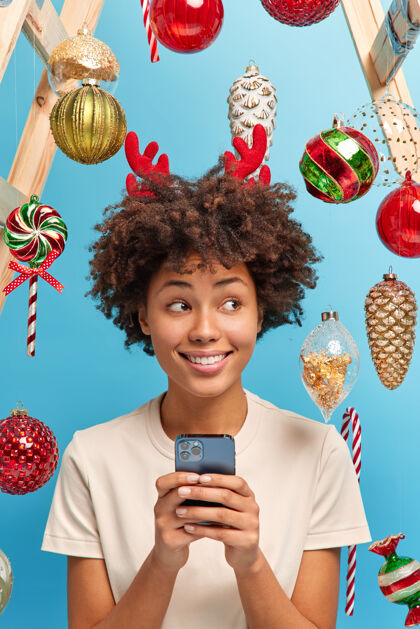 圣诞节空气中弥漫着节日的气息圣诞快乐的概念快乐的黑皮肤女人用智能手机给亲戚们送去祝贺在装饰好的房间里摆出姿势微笑着新年快到了打字女性牙关