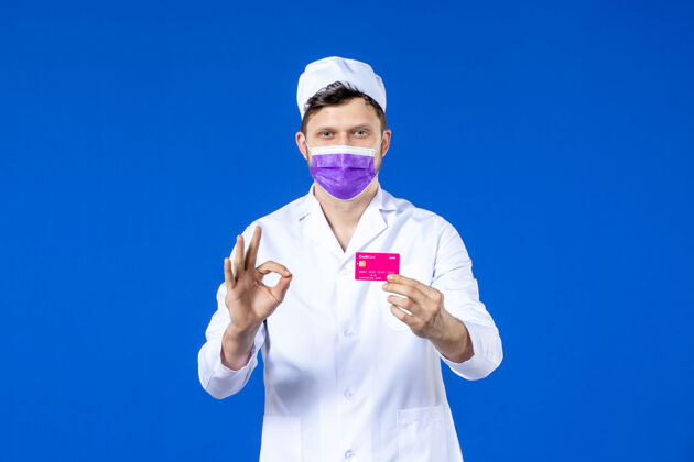 医学前视图中的男医生穿着医疗服和紫色面具持有信用卡在蓝色病毒诉讼大流行