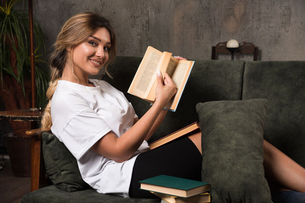 微笑年轻的女人在看一本书 坐在沙发上向前看年轻室内公寓