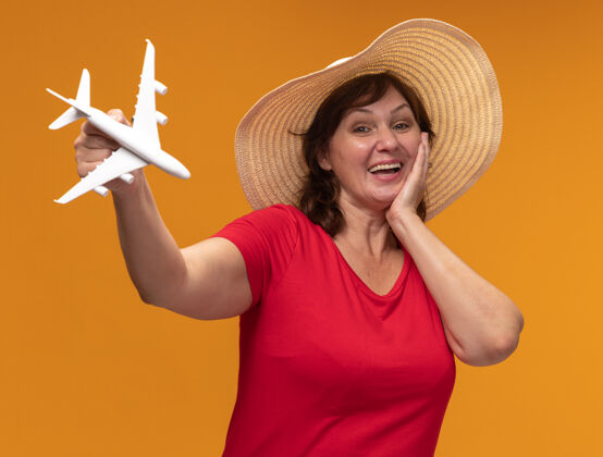 飞机身穿红色t恤 头戴夏帽的中年妇女站在橙色的墙上 脸上露出玩具飞机欢快的笑容中间女人帽子