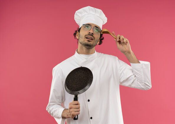 额头看着困惑的年轻男厨师穿着厨师制服 戴着眼镜 拿着煎锅 把勺子放在额头上眼镜困惑薯条