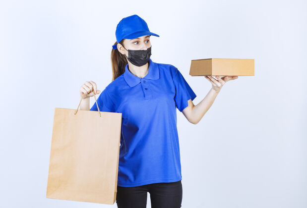 小口带着面具和蓝色制服的女快递员 手里拿着一个纸板购物袋和一个盒子卫生姿势智能