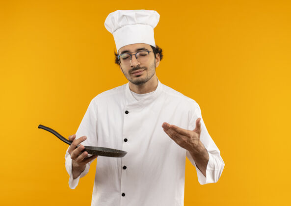 制服年轻的男厨师闭着眼睛 穿着厨师制服 拿着眼镜 用手指着隔离在黄色墙上的煎锅薯条平底锅手