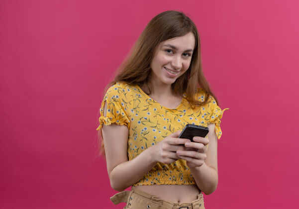 微笑微笑的年轻女孩拿着手机 看着孤立的粉红色墙壁与复制空间手机年轻女人