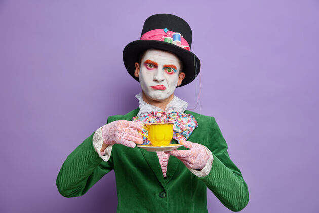 领结无聊的不高兴的男子帽匠沉迷于喝茶戴着大帽子绿色服装蝴蝶结在紫色的墙上摆姿势来参加鬼怪派对庆祝万圣节站在室内射击面具神秘