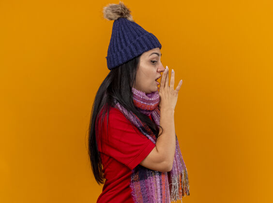 保持印象深刻的年轻白种人生病的女孩戴着冬天的帽子和围巾站在侧视图保持手靠近嘴看边窃窃私语隔离在橙色墙上复制空间近印象疾病