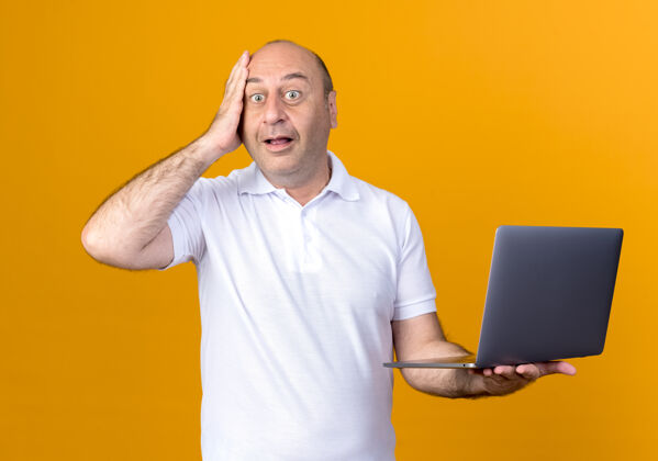 惊喜一个不经意的成熟男人拿着笔记本电脑 把手放在隔离在黄色墙上的额头上笔记本前额手