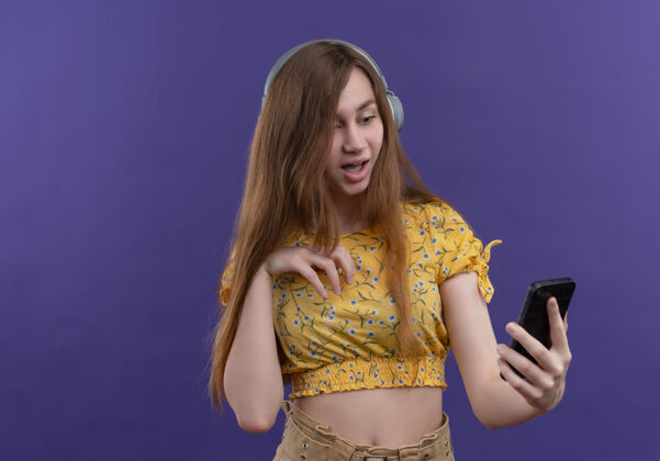 抱惊讶的年轻女孩戴着耳机拿着手机在隔离的紫色墙壁上复制空间女孩耳机手机