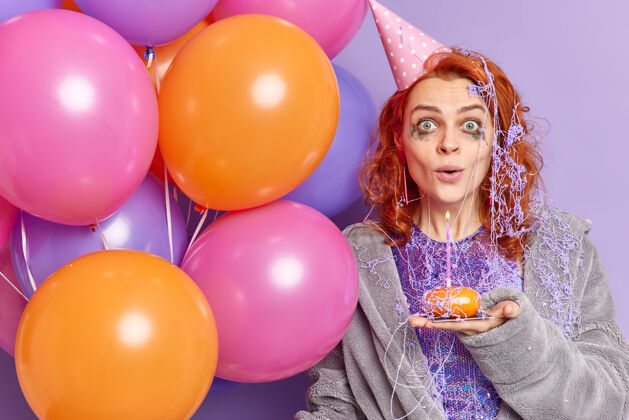 生日在庆祝周年纪念日后 一个被宠坏了妆容的红发女人惊讶地盯着面前 手里拿着纸杯蛋糕和五颜六色的充气气球 孤立在紫色的墙上周年纪念多彩礼物