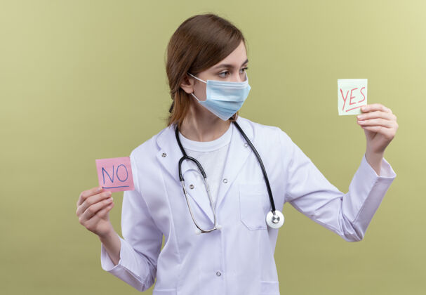 听诊器年轻的女医生穿着医用长袍 戴着口罩和听诊器 拿着是和否的纸条 看着隔离的绿墙上的是纸条年轻女人穿