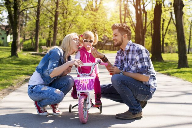 年轻快乐的家庭 年轻的白人父母教他们的女儿如何骑自行车乐趣小休闲