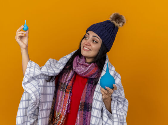 小带着微笑的年轻白种人生病的女孩戴着冬天的帽子 围巾裹着格子布 拿着灌肠器 看着一个被隔离在橙色背景下的小家伙灌肠格子包