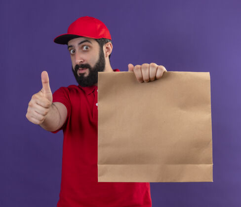 帽子印象深刻的年轻英俊的白人送货员穿着红色制服和帽子伸展纸包对着镜头孤立在紫色红男走向
