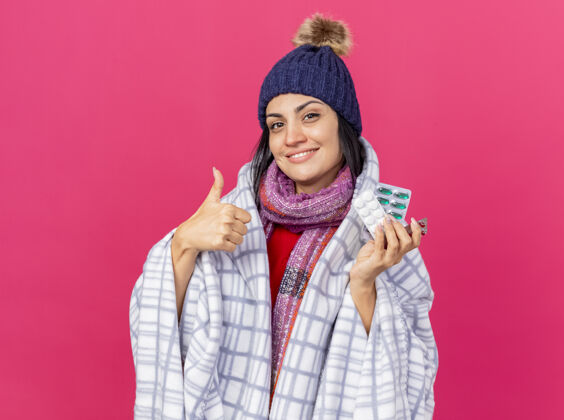 相机带着微笑的年轻白种人生病的女孩戴着冬天的帽子 围巾裹着格子布 看着相机拿着一包包药片 大拇指向上竖起 在深红色的背景上与复制空间隔离开来疾病药片围巾