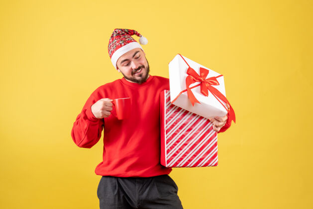 微笑前视图年轻男性 圣诞礼物和黄色背景上的一杯茶礼物背景帽子