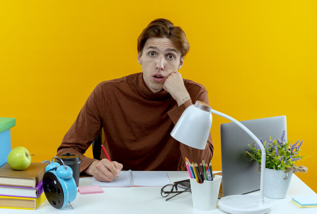 坐着惊讶的年轻学生男孩坐在书桌旁 手里拿着学习工具 在黄色墙上的笔记本上写着什么书桌学校学生