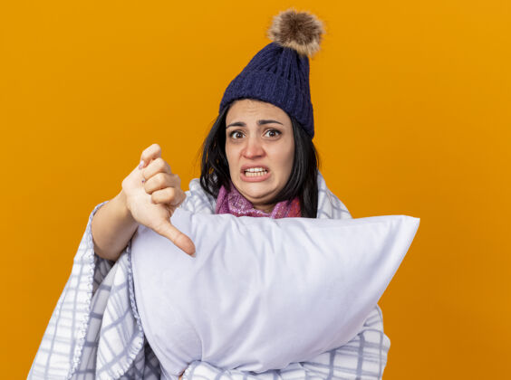 帽子焦急的年轻白种人生病的女孩戴着冬天的帽子 围巾裹在格子布里 抱着枕头看着相机 在橙色的背景下 拇指朝下孤立着裹着戴着焦虑