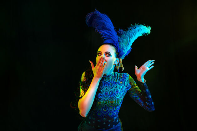 华丽美丽的年轻女子在狂欢节和化装舞会服装在五颜六色的霓虹灯在黑墙上戏剧羽毛节日