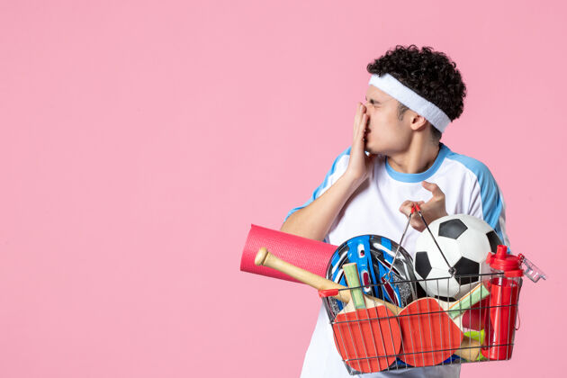 东西正面图穿着运动服的年轻男子 带着装满运动用品的篮子 粉色的墙壁体育商场篮子