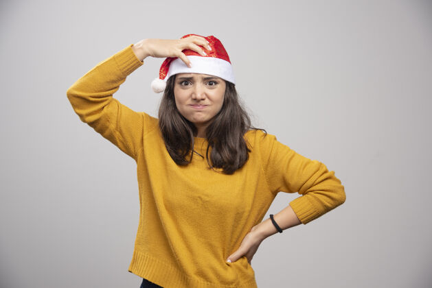 帽子戴着圣诞老人红帽子的年轻女子在白墙上摆姿势圣诞老人女孩模型
