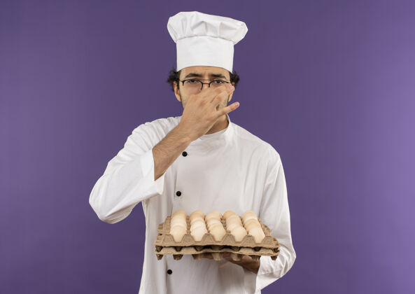 吱吱神经质的年轻男厨师穿着厨师制服 戴着一副眼镜 手里拿着一批鸡蛋 鼻子紧闭在紫色上背景紫色鸡蛋