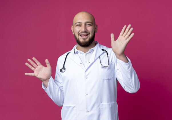 粉色高兴的年轻男医生穿着医用长袍和听诊器把双手分开放在粉红色的皮肤上蔓延手请