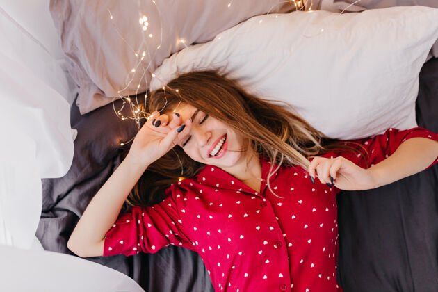 睡衣喜出望外的白人女孩 棕色头发躺在床上 面带微笑穿着红色睡衣的女模特笑着说早上好枕头积极快乐
