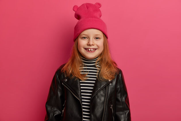 红发时尚正派红发女孩笑容灿烂 缺牙 穿着时尚皮夹克 戴着粉色帽子 享受美好的一天 周末和父母在一起 心情愉快 在室内摆姿势观点服装脸