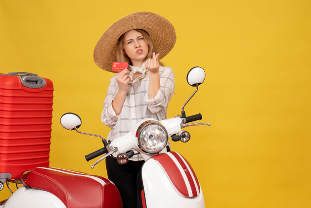 制造正面图是雄心勃勃的年轻女子戴着帽子 坐在摩托车上拿着行李 手里拿着银行卡赚钱的手势钱帽子人