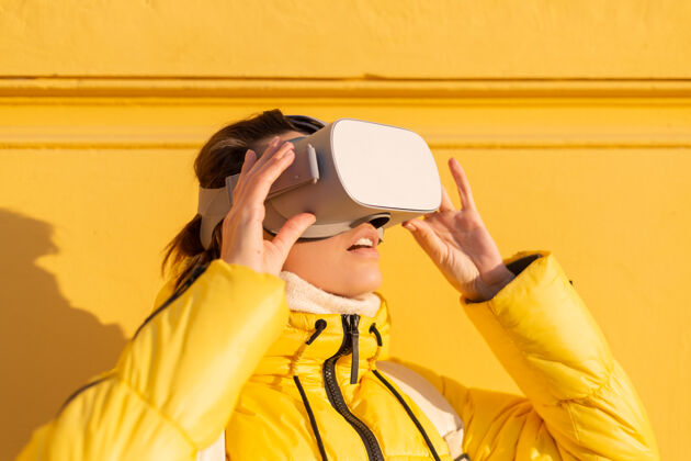 虚拟现实一个戴着虚拟现实眼镜的女人的肖像 在冬天的阳光下 穿着暖和的衣服 靠在黄色的墙上显示玩家数码
