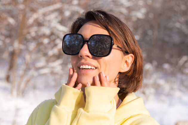 肖像阳光明媚的日子里 雪景冬林中一位年轻美女的画像 身着黄色大套头衫 戴着墨镜 享受着阳光和雪女性微笑白牙