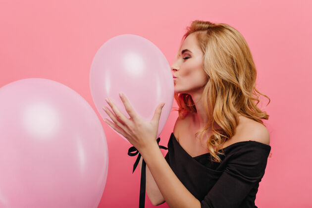 明亮金色头发的幸福女人亲吻粉红色的气球穿着黑色衣服准备活动的令人心寒的年轻女士的室内肖像优雅气球成人