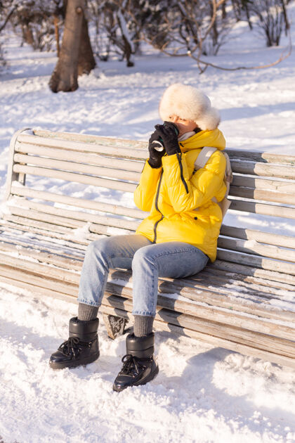 酷冬天的快乐年轻女子 穿着暖和的衣服 在阳光明媚的日子里 坐在长椅上 独自享受着新鲜的空气和咖啡长凳漂亮女孩