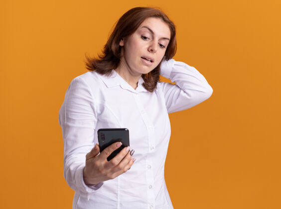 衬衫身穿白衬衫的年轻女子手持智能手机 站在橙色的墙上 看着它困惑不解移动年轻女人