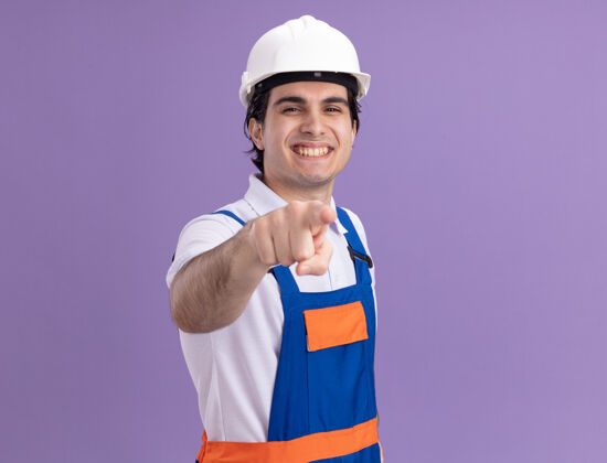 建筑年轻的建筑工人身穿建筑制服 戴着安全帽 食指指着前面 站在紫色的墙壁上 兴高采烈地笑着制服站立手指