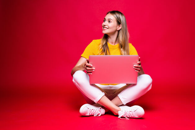 笔记本电脑快乐的年轻卷发美女盘腿坐在地板上 在红墙上用笔记本电脑女孩沟通坐着