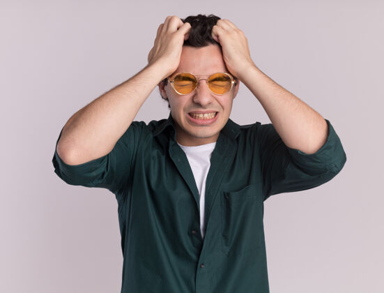 头发一个穿着绿衬衫 戴着眼镜的年轻人站在白墙上狂乱地扯着头发拉着眼镜站着